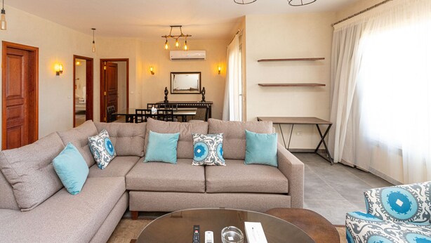 Een woonkamer met een beige bank, twee bijzettafeltjes, een glazen salontafel, en een eethoek met een tafel en stoelen.