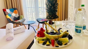 A hotel room with a dining table set with a fruit platter, a pineapple centerpiece, and a bottle of water. There is a chair with a colorful cushion and a towel with the hotel's logo. - Vinea Resort & SPA (Rrashbull)
