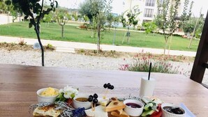 A wooden table with a variety of food items including cheese, olives, and bread, set outdoors with a view of a landscaped garden and buildings in the background. - Vinea Resort & SPA (Rrashbull)