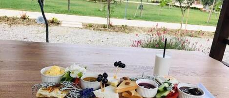 Una mesa de madera con diversos alimentos, como queso, aceitunas y pan, ubicada al aire libre con vistas a un jardín ajardinado y edificios al fondo.