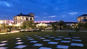 A well-lit outdoor area with a checkered pattern of grass and concrete, surrounded by buildings with illuminated windows. - Vinea Resort & SPA (Rrashbull)