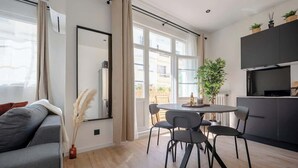 A modern living room with a grey sofa, a round dining table with chairs, a large window with curtains, and a kitchen area with cabinets and a countertop.