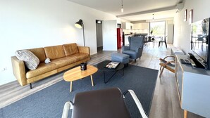Ein modernes Wohnzimmer mit Sofa, Sessel, Couchtisch und Fernseher. Im Hintergrund ist eine Küche zu sehen.