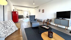 Ein modernes Wohnzimmer mit Sofa, Sessel, Couchtisch und Fernseher. Im Hintergrund sind ein roter Kühlschrank und ein Essbereich zu sehen.