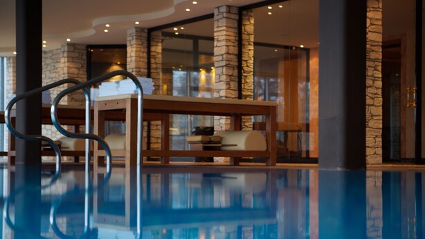 Un área de piscina interior moderna con columnas de piedra, un banco de madera y una barandilla de cristal.