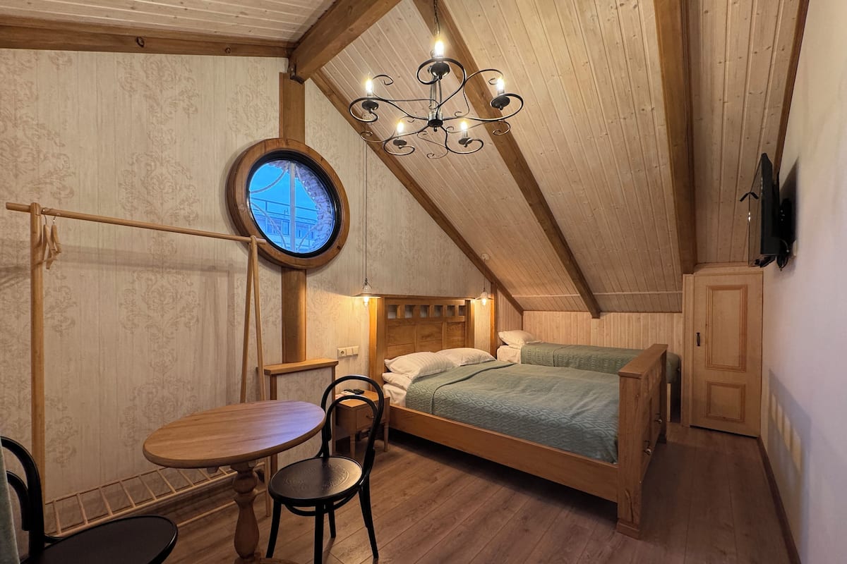 A cozy attic bedroom with a wooden ceiling, a round window, a bed, a table, and chairs.