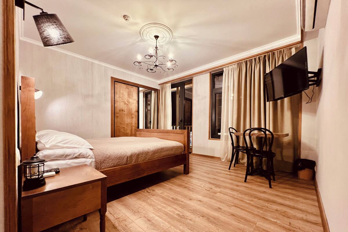 A bedroom with a wooden bed, a nightstand, a desk, a chair, a TV mounted on the wall, and a chandelier.