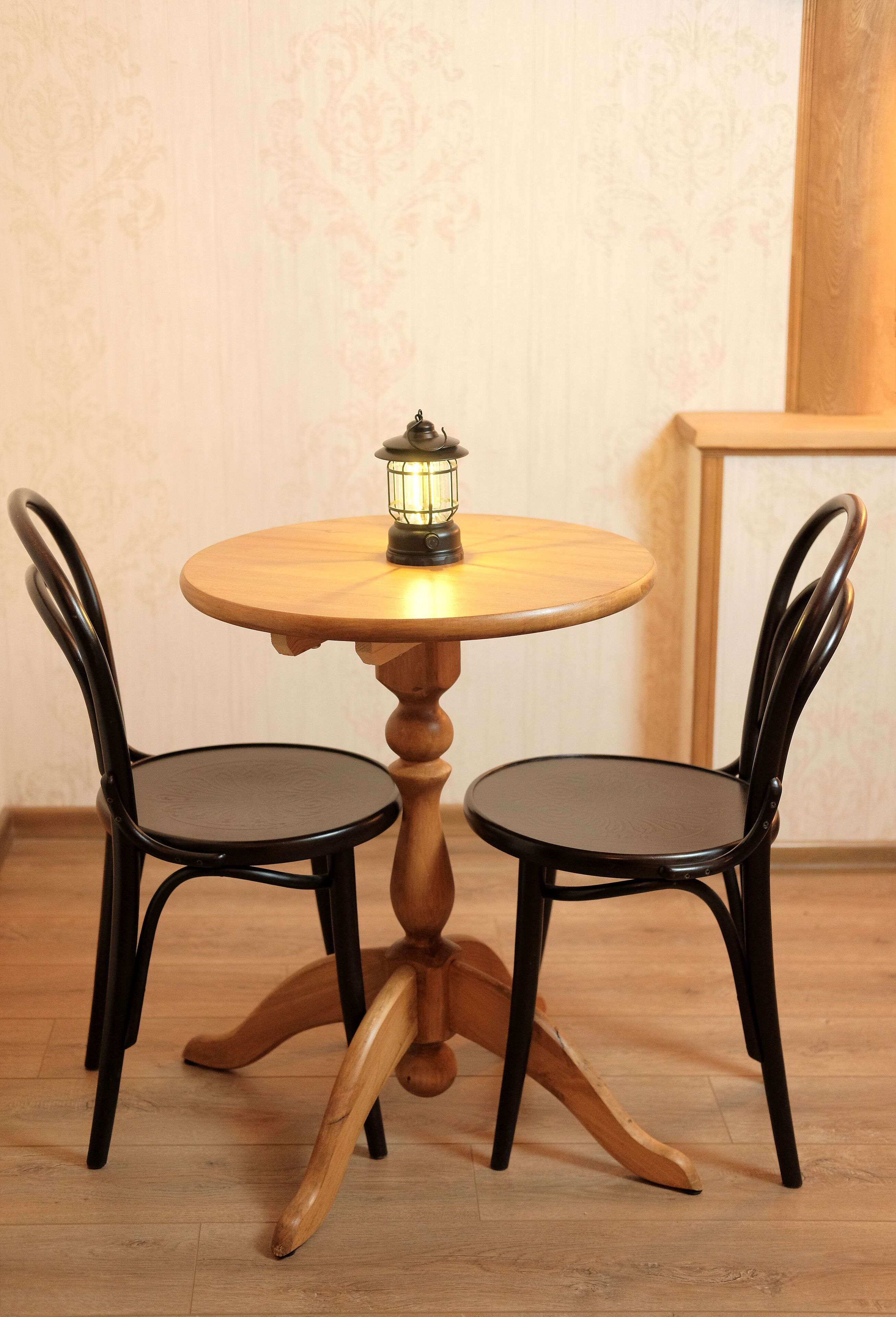 Ein runder Holztisch mit einer einzelnen Kerze, daneben zwei schwarze Stühle mit geschwungenen Rücken.