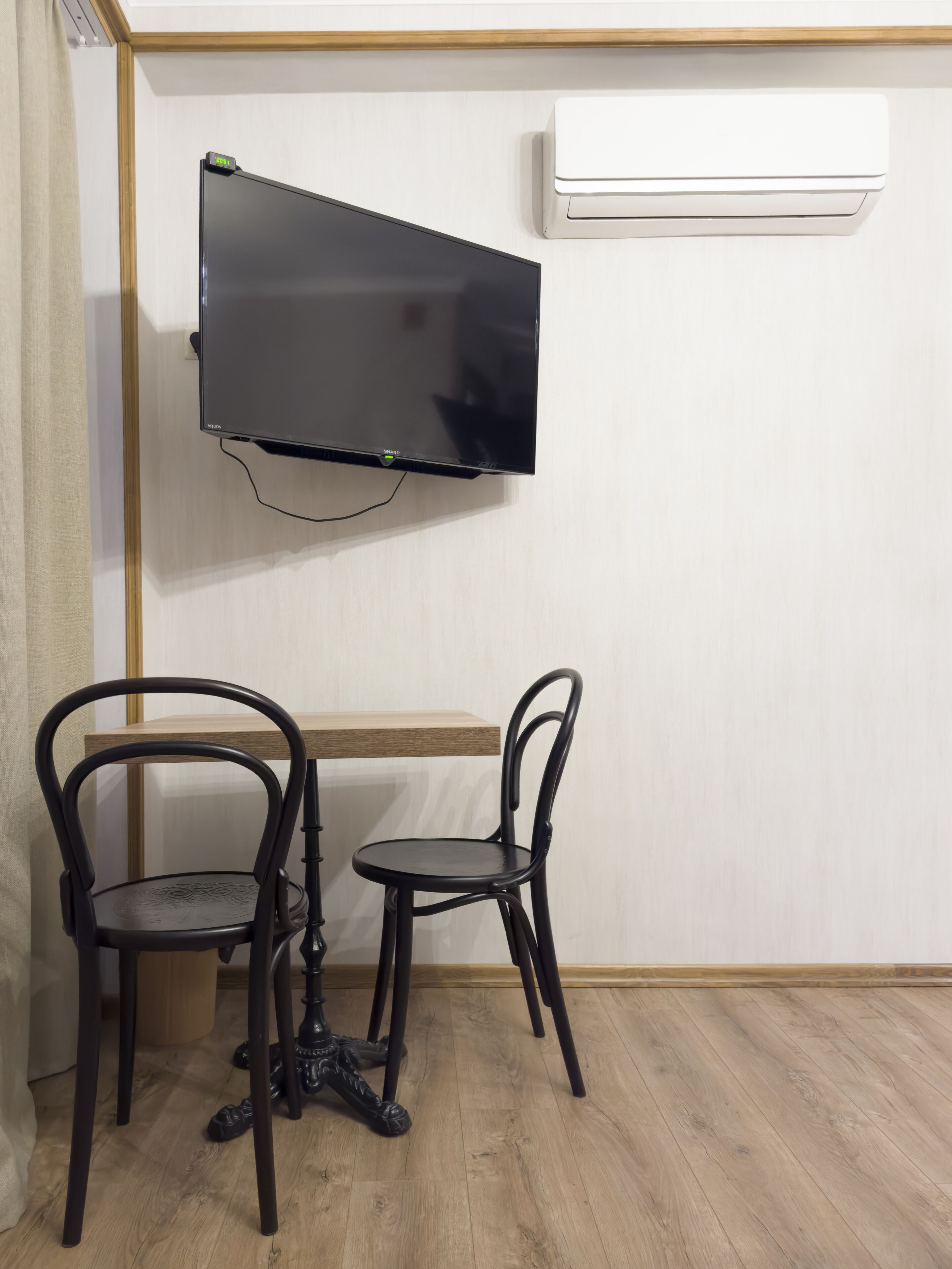A room with a flat-screen TV mounted on the wall, a wooden table, two black chairs, and a white air conditioning unit.