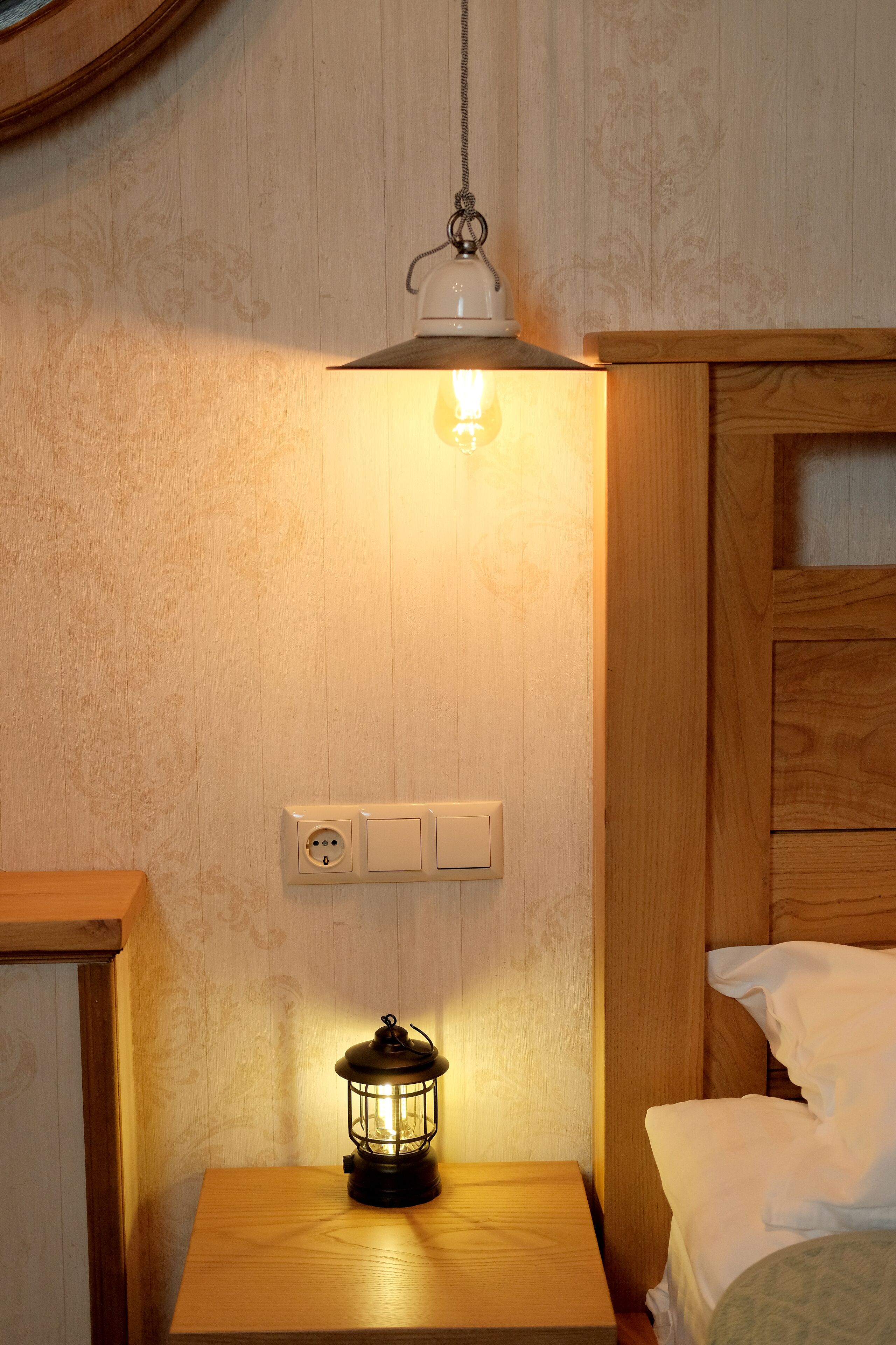 Una mesita de noche de madera con un farol, un enchufe de pared y una cama con ropa blanca.