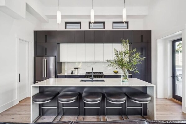 Una cocina moderna con una isla central, taburetes de bar, armarios oscuros e iluminación colgante.