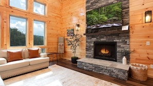 A cozy log cabin interior with a stone fireplace, a sofa, and a large window overlooking a forest. - Mountain Views/Near DwTn Gat/New (Gatlinburg)