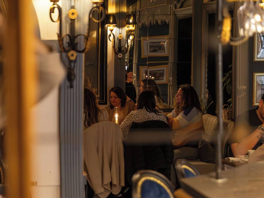A room with a mirror reflecting a group of people sitting on a couch. There are wall-mounted lamps and a chandelier visible.