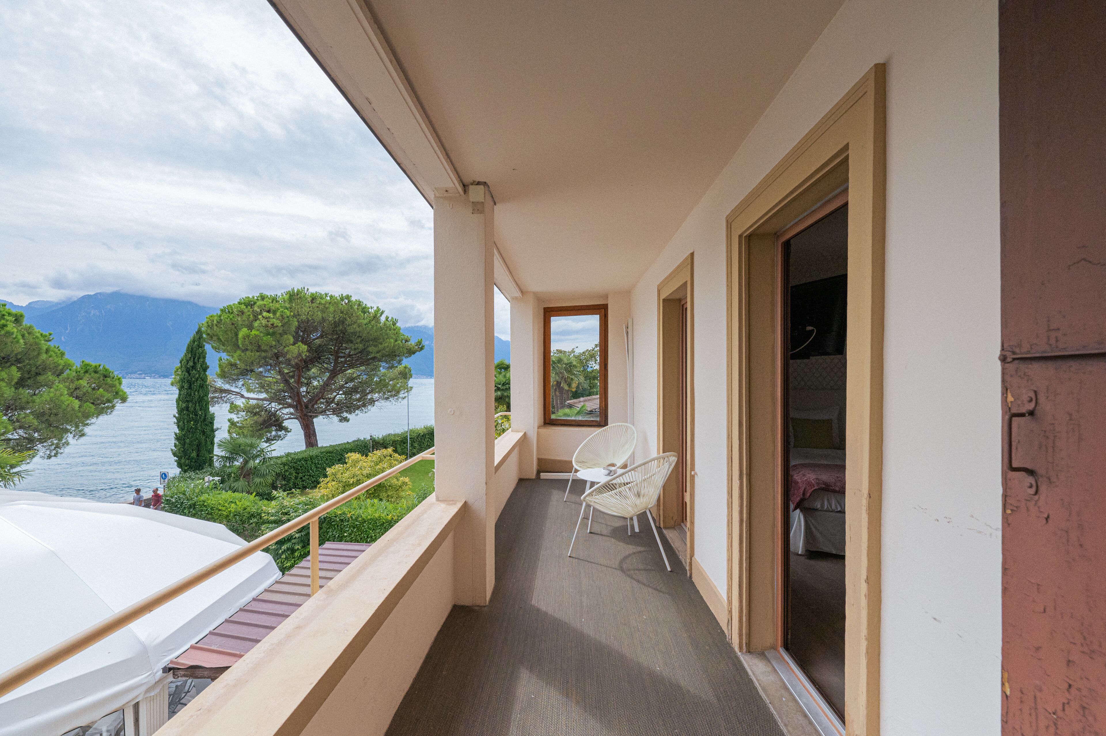 A balcony with a view of a lake and mountains, a white chair, and a door leading to a room.