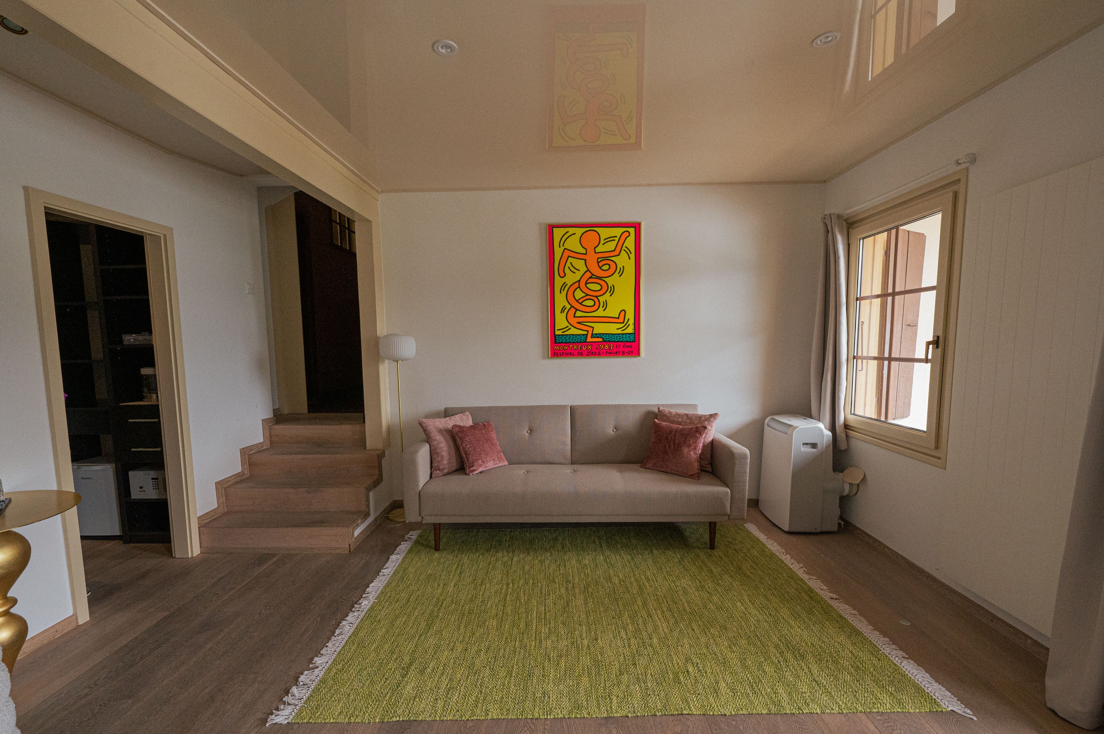 Un salon avec un canapé beige, un tapis vert, un tableau au mur et une fenêtre avec des rideaux.