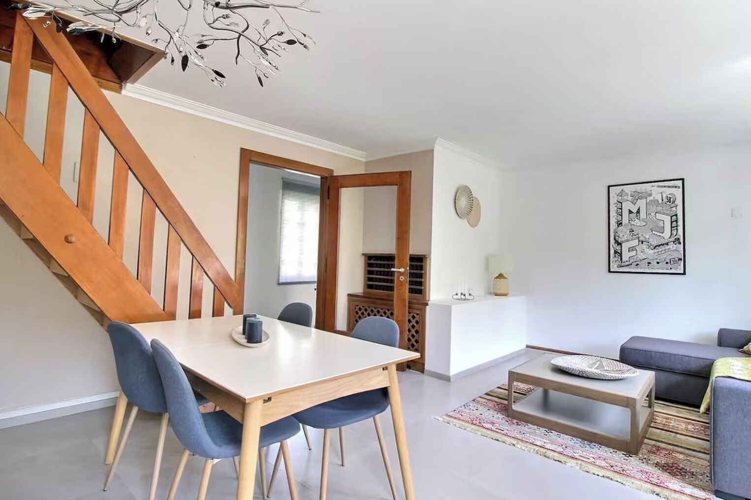 Un espacio de estar moderno con una escalera de madera, una zona de comedor con mesa y sillas, un sofá y una mesa auxiliar.