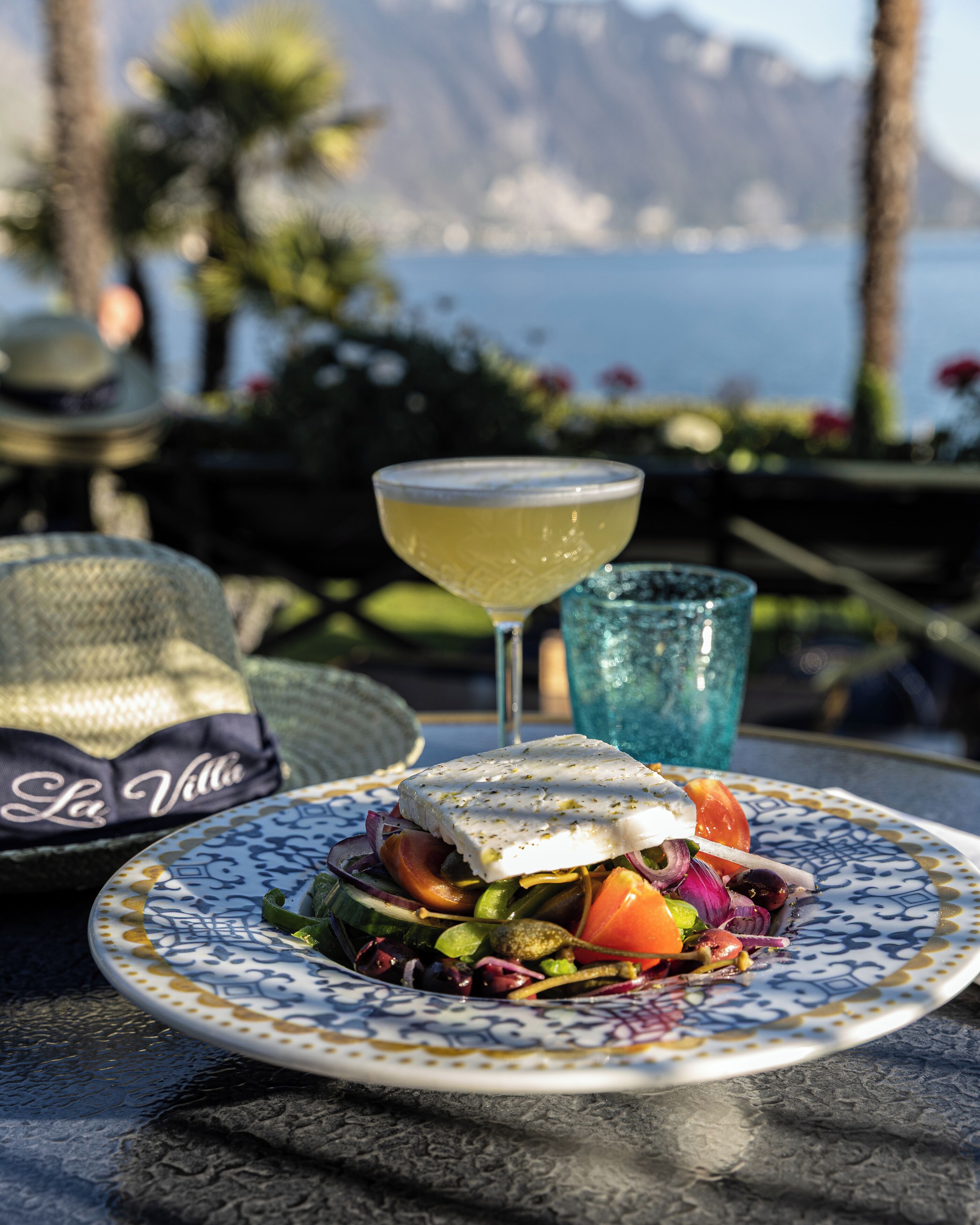 A meal with a fish dish and salad on a patterned plate, a cocktail glass, and a hat with "La Villa" on it.