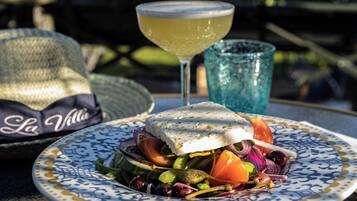 A meal with a fish dish and salad on a patterned plate, a cocktail glass, and a hat with "La Villa" on it.