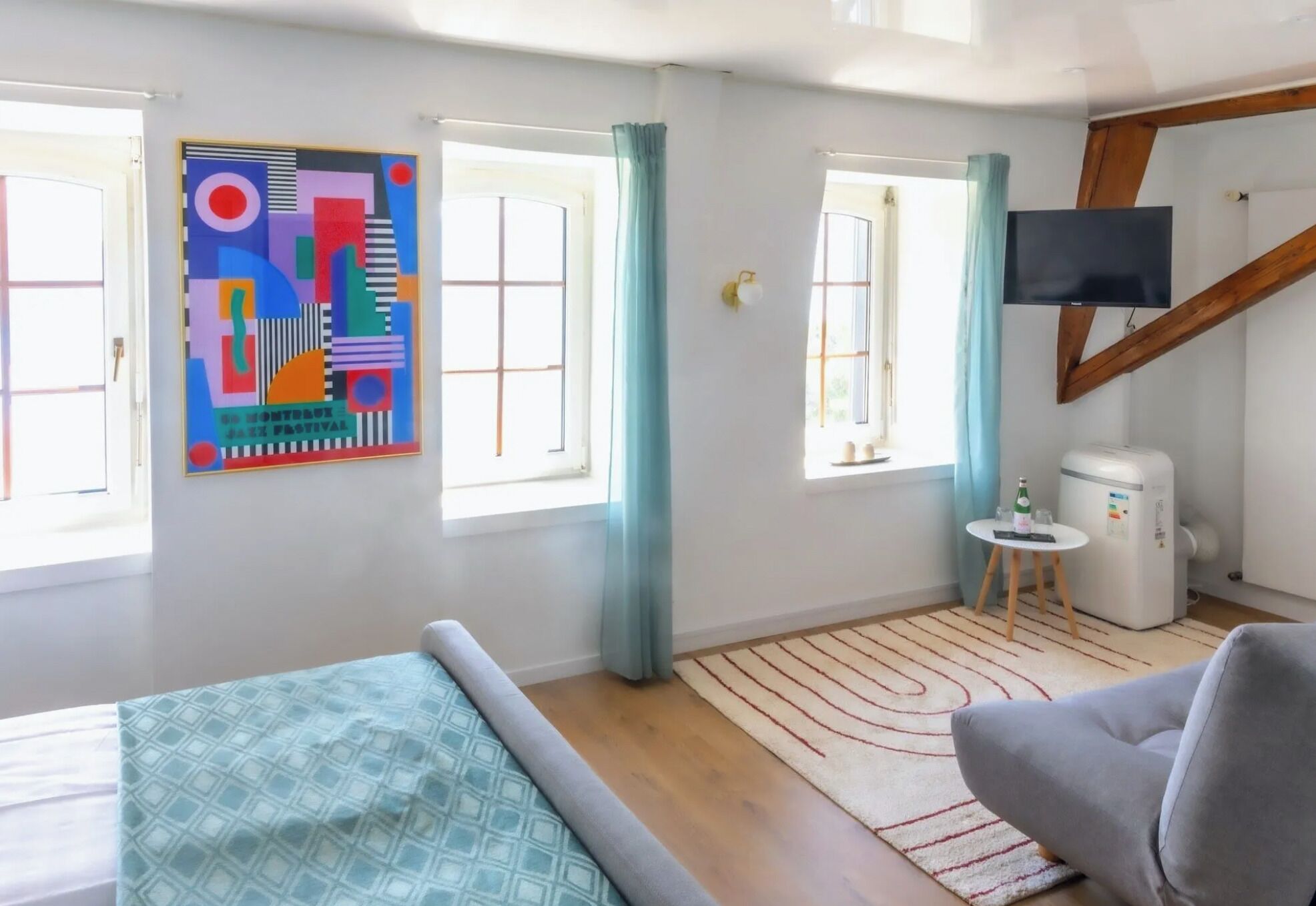 Ein modernes Zimmer mit einem Bett, einem grauen Sofa, einem kleinen Tisch mit einer Flasche, einem Fernseher und einem farbenfrohen, abstrakten Gemälde an der Wand.