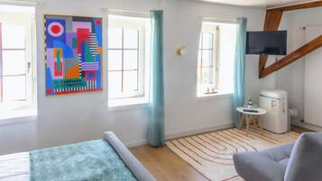 Ein modernes Zimmer mit einem Bett, einem grauen Sofa, einem kleinen Tisch mit einer Flasche, einem Fernseher und einem farbenfrohen, abstrakten Gemälde an der Wand.