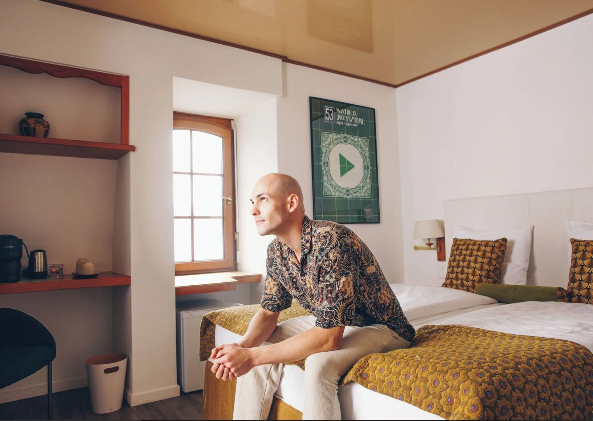 A hotel room with a bed, a desk, a chair, a shelf with items, and a window.