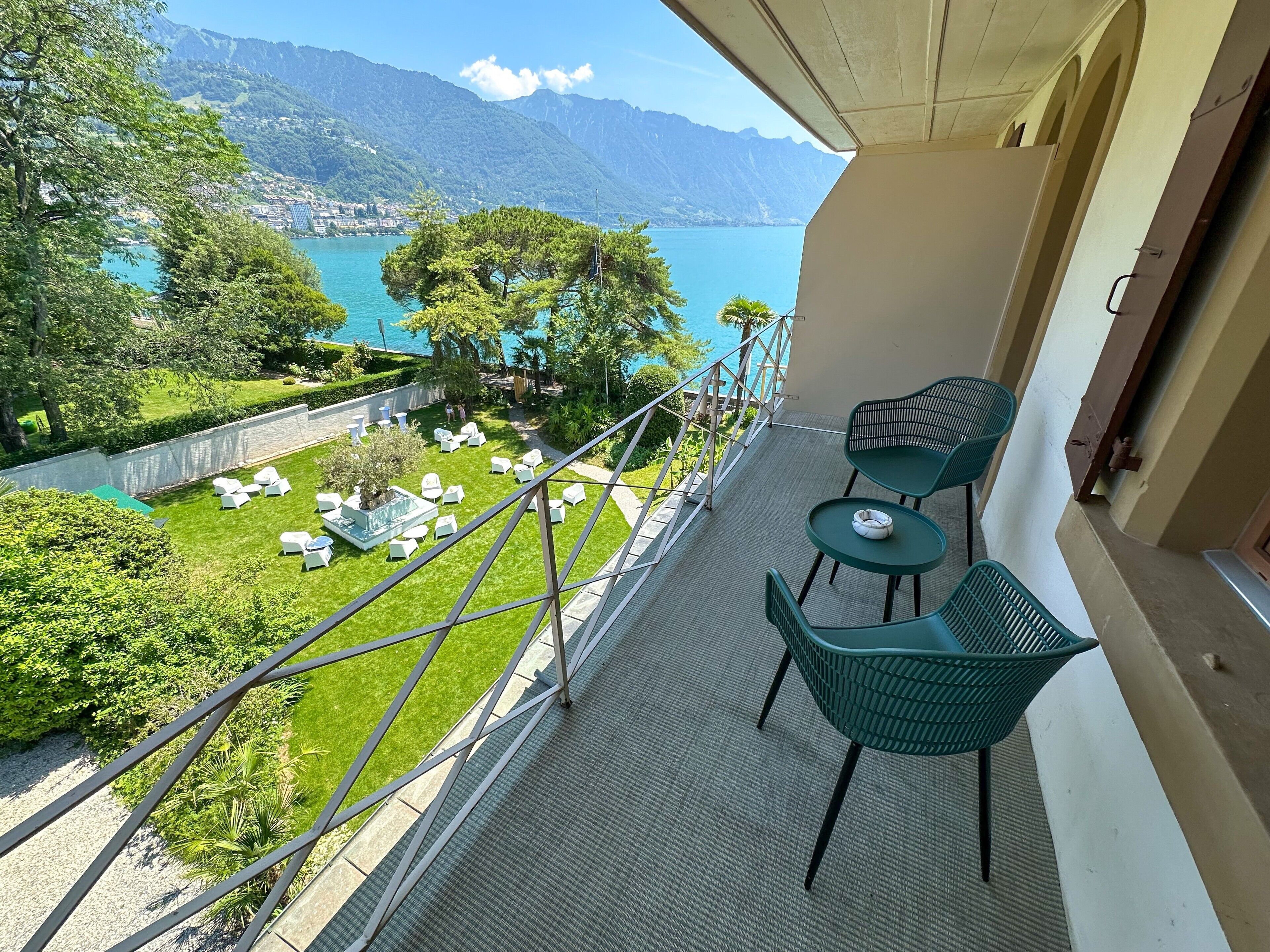 Un balcon avec vue sur un lac et des montagnes, des chaises vertes et une petite table.