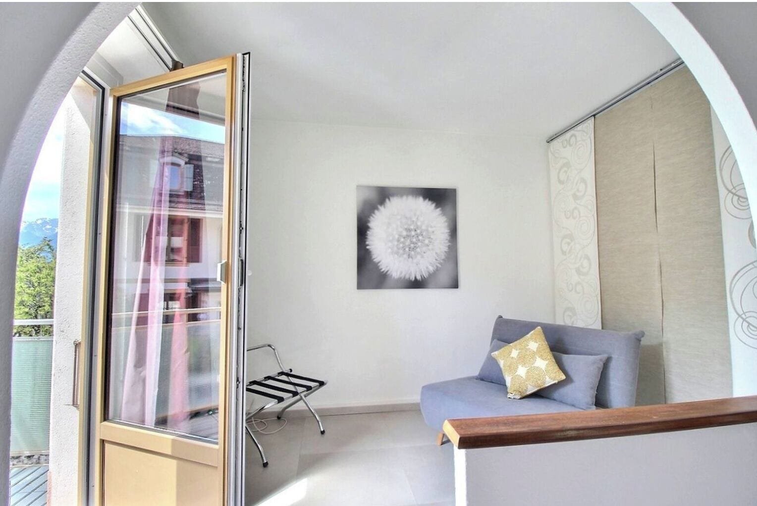 A room with a sliding glass door leading to a balcony, a grey sofa with a patterned cushion, a wall-mounted artwork, and a small black drying rack.