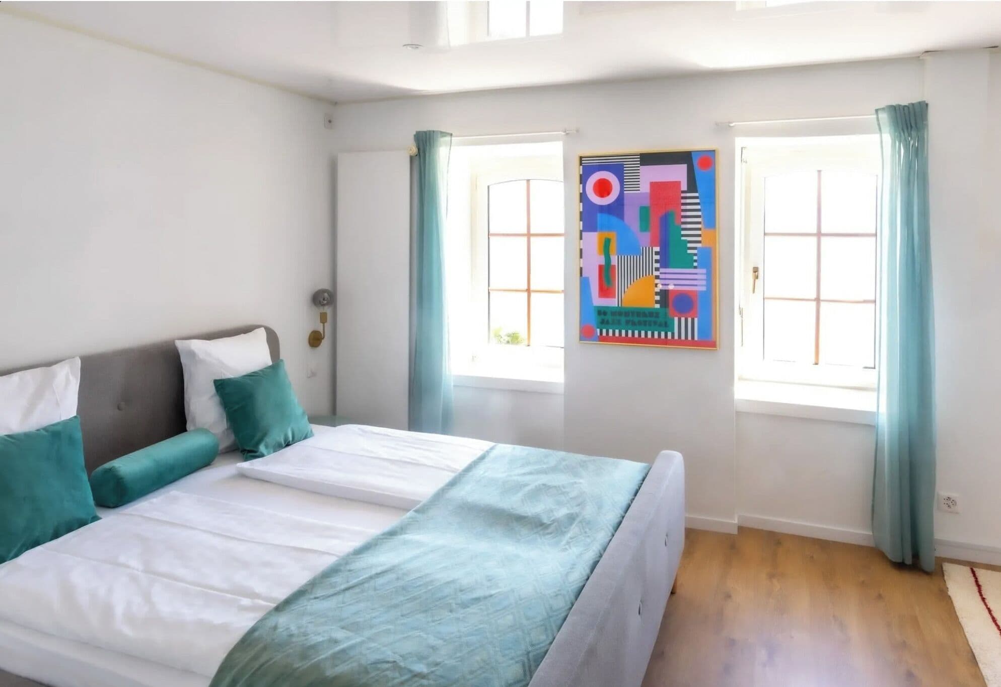 A bedroom with a bed, two windows with curtains, and a colorful abstract painting on the wall.
