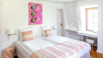 Una cama doble con colcha a rayas, una mesita de noche con lámpara, un cuadro de pared rosa, y una ventana con vistas al exterior.