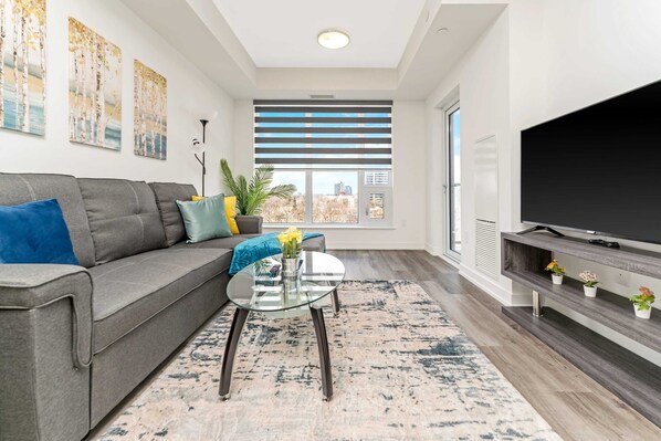 A modern living room with a grey sofa, a glass coffee table, a TV on a wooden stand, and a large window with blinds. - Newly Built Modern Condo w Balcony & City Views (Kitchener)