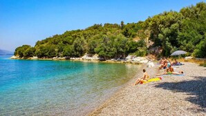 A pebble beach with clear turquoise water, people relaxing, and a forested hill in the background. - Meganisi Villa Armonia - Private Pool Oasis (Vathi)