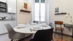 A dining area with a round table set for four, surrounded by modern chairs. There is a kitchen area to the left with a shelf above the counter. - Italianway - Ozieri 7 (Milan)