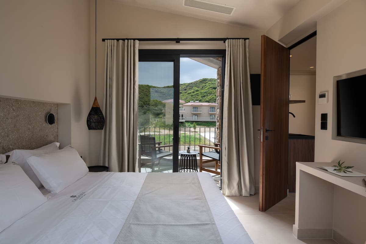 A hotel room with a large bed, a desk, a TV, and a balcony with a view of the hills.