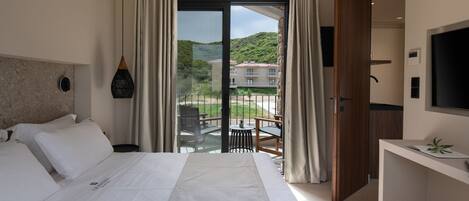 A hotel room with a large bed, a desk, a TV, and a balcony with a view of the hills.