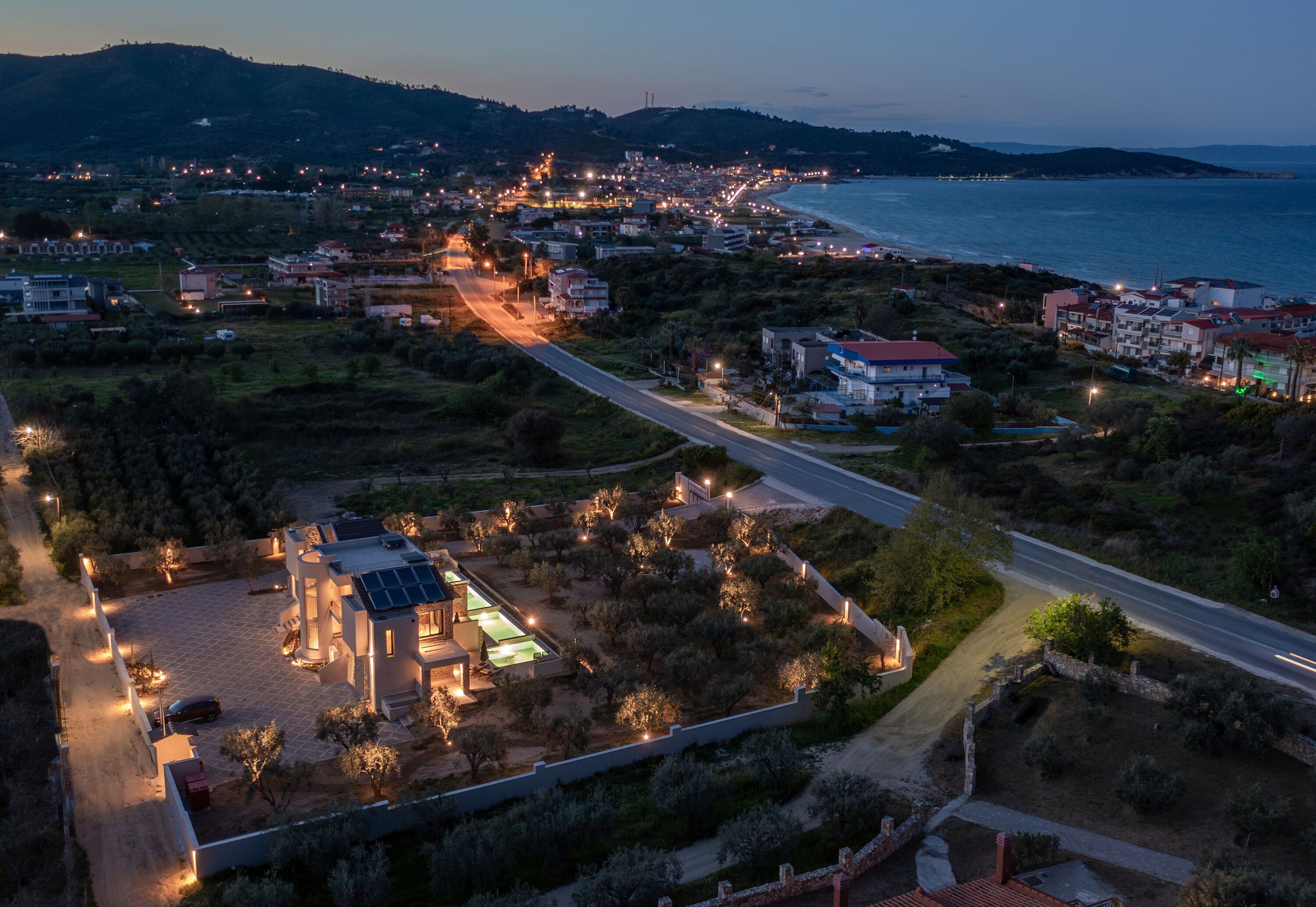 An aerial view of a coastal town with a prominent road, residential buildings, and a large property with a solar panel array.