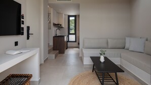 A modern living room with a sofa, a coffee table, and a TV on a white wall. There is a kitchen area with a sink and cabinets in the background.