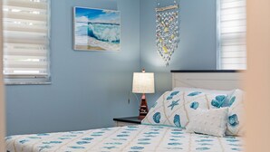 A bedroom with a bed, a nightstand, a lamp, a wall hanging, and a window with blinds. - Coastal Breeze on 66th Avenue (Pinellas Park)