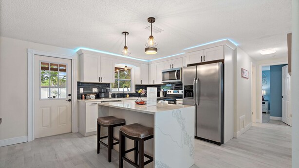 A modern kitchen with white cabinets, stainless steel appliances, and a marble island with bar stools. - Coastal Breeze on 66th Avenue (Pinellas Park)