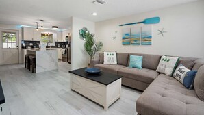 A modern kitchen with a central island, a dining area with a sofa and cushions, and a living room with a coffee table. - Coastal Breeze on 66th Avenue (Pinellas Park)
