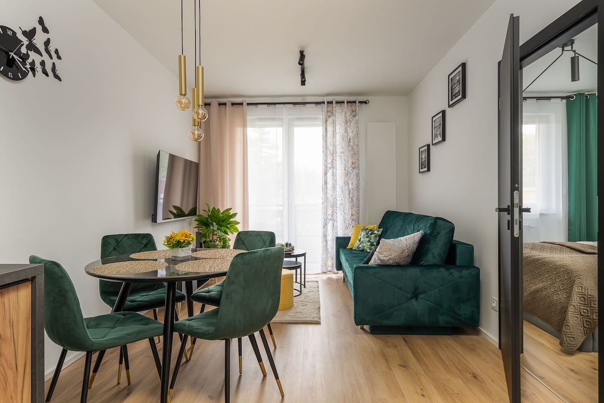 A modern living room with a green sofa, a dining table with chairs, a TV mounted on the wall, and a large window with curtains.