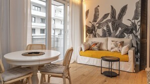 A modern living room with a round dining table, wicker chairs, a sofa with a yellow throw, and a large window with a view of a building. - RentPlanet - Apartament Długa (Wroclaw)