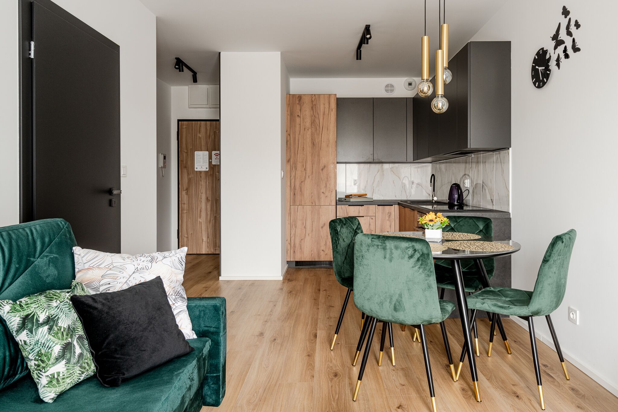 A modern kitchen with dark cabinets, a wooden door, and a dining area with green velvet chairs and a round table.