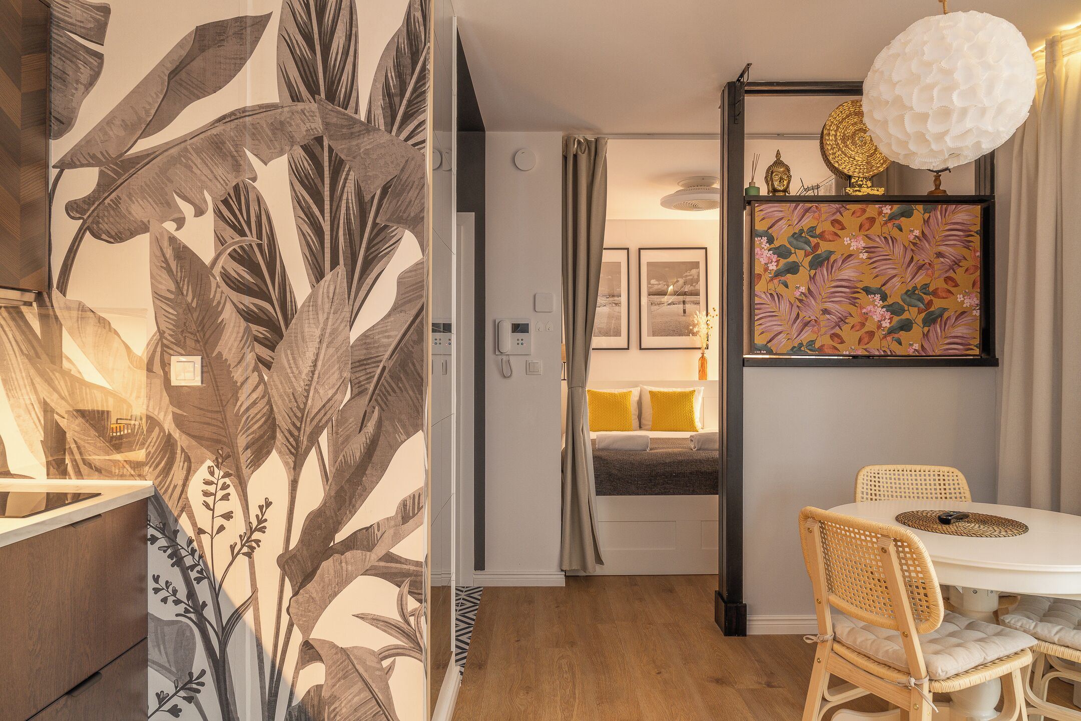 A room with a bed, a dining table with chairs, a wall with a tropical leaf pattern, and a hanging light fixture.