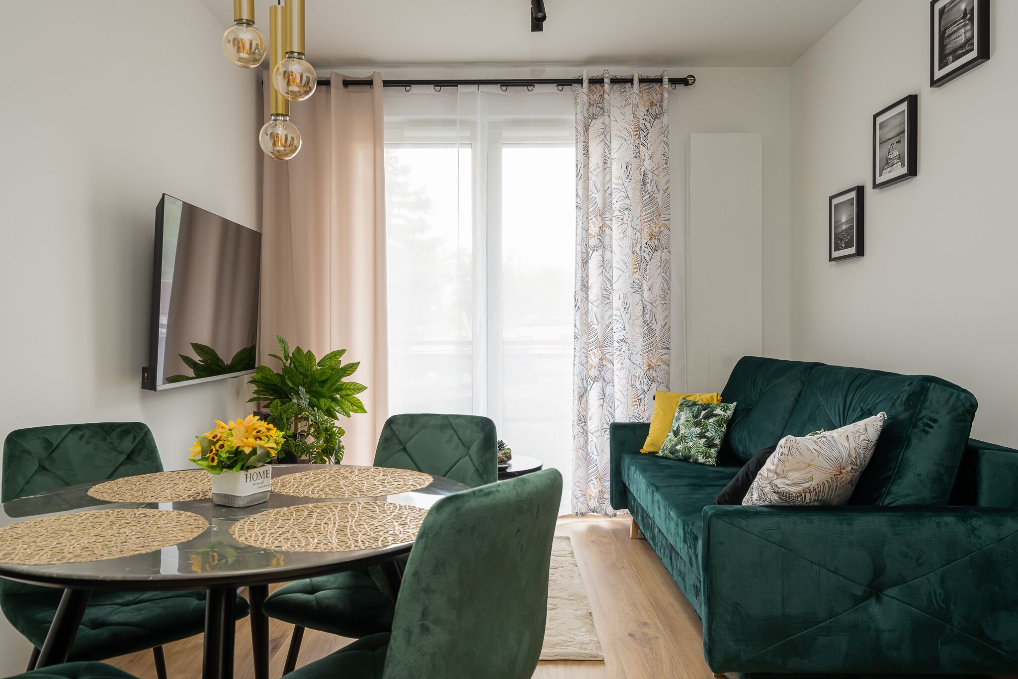 A modern living room with a green sofa, a dining table with four chairs, and a TV mounted on the wall.