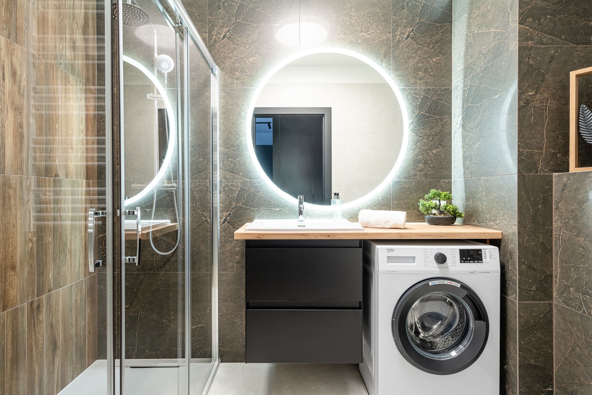 A modern bathroom with a glass shower enclosure, a round mirror, a sink with a wooden countertop, and a front-loading washing machine.