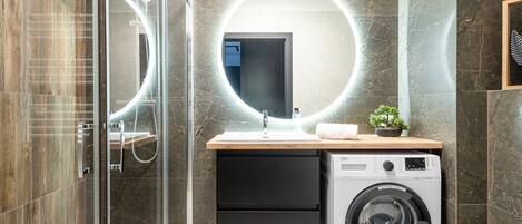 Un baño moderno con cabina de ducha de vidrio, un espejo redondo, un lavabo con encimera de madera y una lavadora de carga frontal.