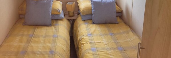 Two single beds with yellow plaid bedding and blue pillows, a bedside table with a lamp, and a wooden door. - Luxury 8 berth spacious caravan (Chapel Saint Leonards)
