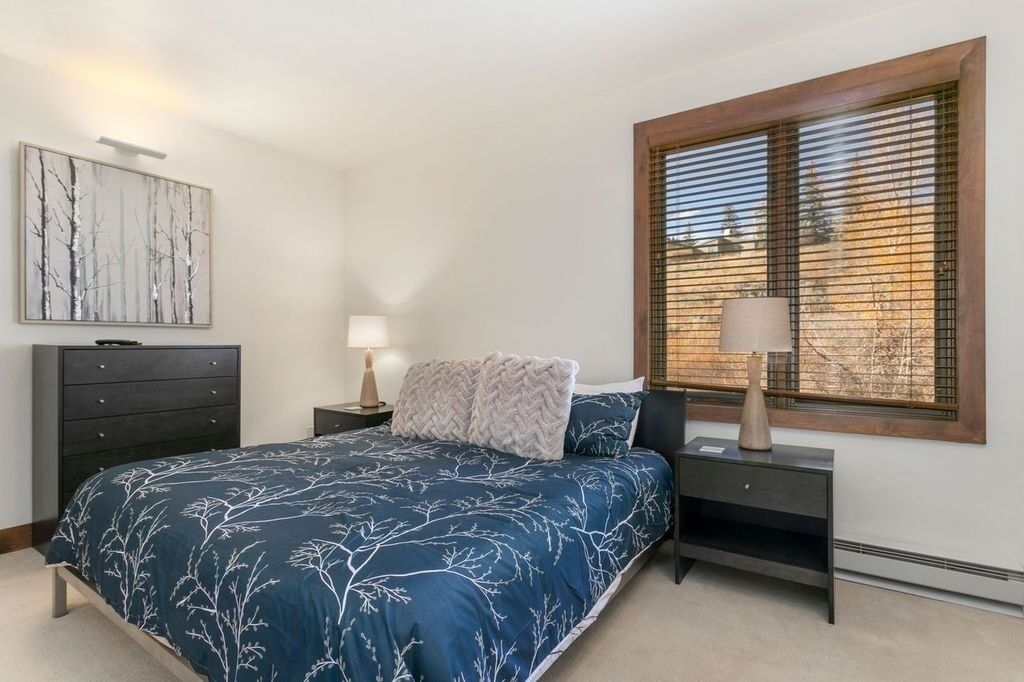 A bedroom with a bed, a nightstand, a lamp, a dresser, a window with blinds, and a wall-mounted artwork.