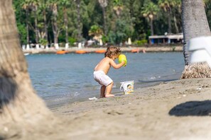 A child playing with a bucket on a sandy beach with a calm body of water and trees in the background. - Mosso Boutique Hotel (Ula)