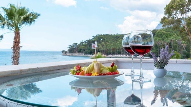 A glass table with a fruit platter and two wine glasses, set against a scenic coastal backdrop. - Mosso Boutique Hotel (Ula)